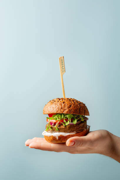 Hand holding burger. Hamburger with meat free plant based cutlet, tomatoes, onions, white sauce and microgreens on blue background. Healthy vegan or vegetarian food concept. Copy space. Hand holding burger. Hamburger with meat free plant based cutlet, tomatoes, onions, white sauce and microgreens on blue background. Healthy vegan or vegetarian food concept. Copy space. veggie burger stock pictures, royalty-free photos & images