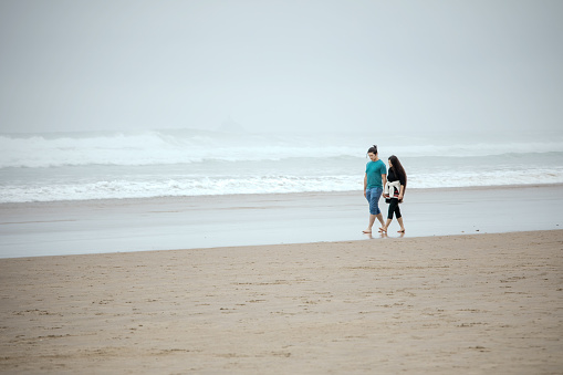 Biracial young couple in twenties walking on empty beach next to ocean on foggy day