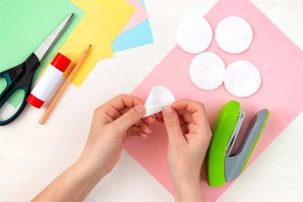 istock Women's hands make a petal from cotton wool. Colored paper, scissors, scissors, stapler, pencil on a white table. Children's crafts for mother's day. Step-by-step instruction. Step 2. 1313817624