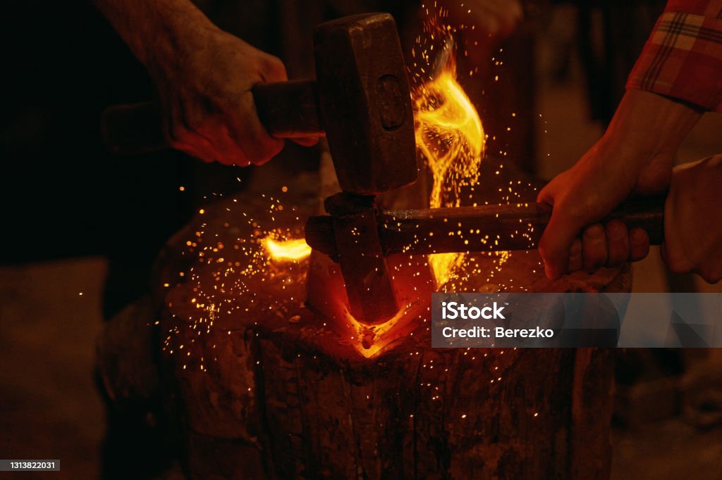 Blacksmiths hit molten metal with hammers close up Blacksmiths forging in smithy. Close up of hands of craftsmen hit molten metal detail with hammers into burning fire on wooden timber. Handmade metal work idea. Concept of handicraft Blacksmith Stock Photo