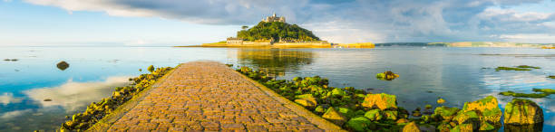 St Michaels Mount sunrise over calm sea panorama Marazion Cornwall Gentle ocean tide ebbing across the iconic cobbled causeway as warm summer sunlight illuminates the historic chapel, hamlet and harbour of St. Michael's Mount, Marazion, Cornwall, UK. marazion photos stock pictures, royalty-free photos & images