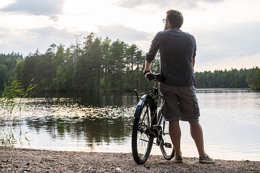 Man with a bicycle stopped on the shore of a forest lake and enjoys the sunset and the beautiful landscape on a cloudy, summer day.