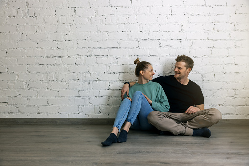 young happy couple sitting on the floor by the wall in empty apartment room. moving to new home concept. copy space