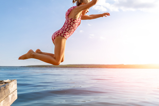 Little cute kid girl in swimsuit have fun enjoy pretend flying jumping from pier dock in clean blue water sea river or ocean on hot summer evening sunset. Carefree children lifestyle vacation concept.