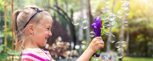 Portrait of cute little bond kid girl enjoy having fun play blowing soap bubbles at home yard garden outdoors on bright warm summer day against lawn and trees. Child healthy outside nature activities. retrato de linda niña bond niña disfrutar de divertirse jugar soplando burbujas de jabón en el jardín del patio de casa al aire libre en brillante día de verano cálido contra el césped y los árboles. actividades infantiles saludables fuera de la na