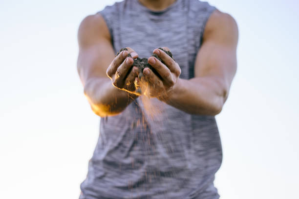 A farmer wearing a blue shirt with the sleeves rolled up and leather gloves holding a potato in his hand as the dirt falls from the freshly dug up potato against a black background 