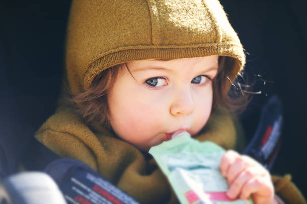Close up portrait of sweet toddler kid eating fruit puree from plastic doy pack, sitting in stroller, outdoor snack time stock photo