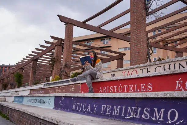Photo of View from below of a woman reading a book, sitting on bleachers that look like books. One can read titles such as ethics and feminism, tao, Aristotle ...