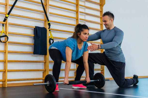 par de um personal trainer sorridente e uma linda mulher grávida fazendo exercício de treino dentro de casa.menina levantando peso em uma sessão de ginástica dentro. fit, treinamento, fitness, conceito de saúde e amizade - crouching barbell weightlifting weight training - fotografias e filmes do acervo