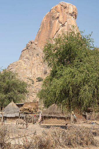 Typical Chadian village in front of the rock tower Pic de Guera (Guera Massiv). \n\nThe province of Guéra, located in central Chad, about 500 km from the capital N'Djamena, is settled by cattle breeders and agricultural farmers, who live in a synergic relationship in which the mountains are sacred and are home to the Margay, the ancestor spirits of the Animist culture.