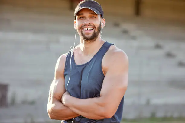 Photo of Shot of a sporty young man wearing his earphones and standing with his arms crossed outside