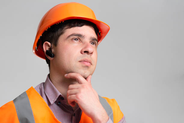 istock Young engineer architect in hard hat and reflective vest thinking with hand under chin on grey studio background, building industry concept 1313893735