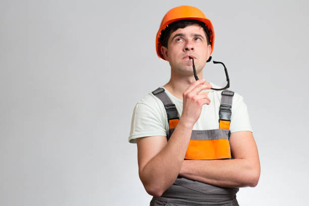 istock Young builder constructor in hard hat and overalls thinking with glasses in mouth on studio background, building industry concept 1313893768