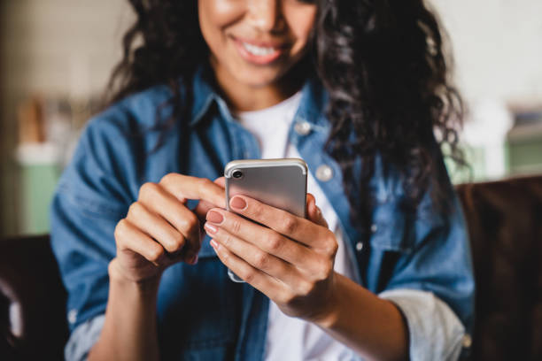 foto recortada de una joven afroamericana usando un teléfono inteligente en casa. mujer afroamericana sonriente usando teléfono inteligente en casa, mensajería o navegando por las redes sociales mientras se relaja en el sofá - una sola mujer fotografías e imágenes de stock