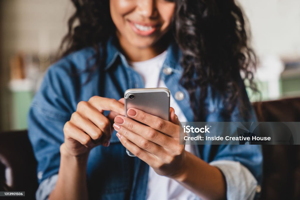 一個非洲裔美國年輕女子在家使用智慧手機的鏡頭。微笑的非洲裔美國婦女在家裡使用智能手機， 在沙發上放鬆時發短信或流覽社交網路 - 免版稅使用電話圖庫照片