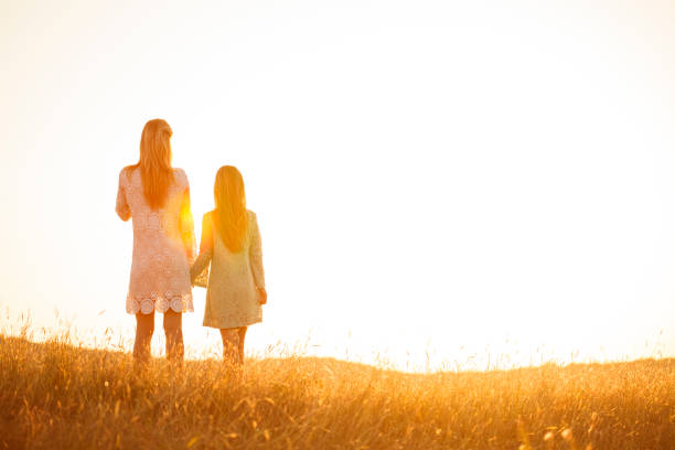 Wide shot of mother and her daughter standing in a golden meadow, holding hands and enjoying watching the beautiful summer sunset together.