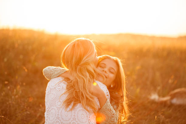 Candid shot of loving mother embracing her young daughter and sharing a moment of affection with her during a golden hour sunset in the meadow.