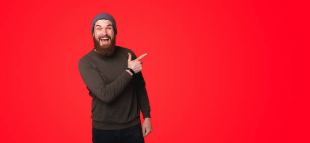 istock An excited young bearded man while he is smiling at the camera is pointing at a free blank space on a red wall 1313902195