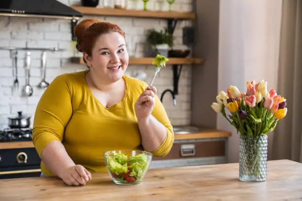 Photo of Obese young caucasian woman eating salad for healthy diet and eating habits in the kitchen. Chubby plus size woman having salad for breakfast