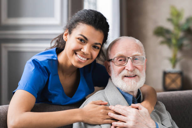 freundlicher reifer allgemeinmediziner kommuniziert mit angenehmen 80er jahren männlichen patienten, sitzen zusammen auf dem sofa. lächelnd vertrauensvolle junge arzt, der psychologische hilfe für älteren mann zu hause besuch. - social worker stock-fotos und bilder