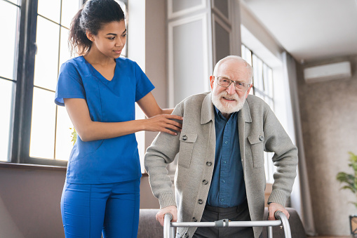 Handicapped man using walker - rehabilitation at home. Nurse helping disabled man using walker in rehab center. Happy female nurse helping elderly grandfather to walk using walker in hospital. Doctor woman giving physical therapy to senior man.