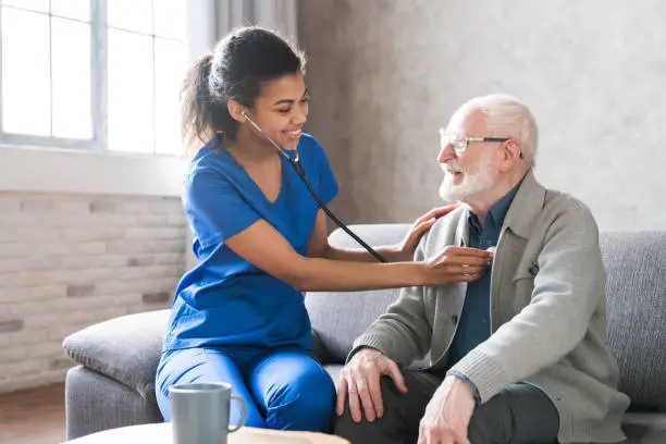 Female attending physician holding stethoscope listening old patient during homecare visit. Doctor checking heartbeat examining elder retired man at home. Seniors heart diseases, cardiology concept.