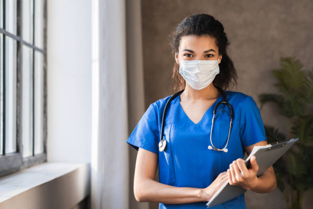 la joven enfermera africana de ropa africana, segura de sí misma, usa uniforme azul, máscara facial, brazos cruzados en el pasillo del hospital. negro millennial mujer doctora, cirujano, médico personal retrato profesional. - medical assistant fotografías e imágenes de stock