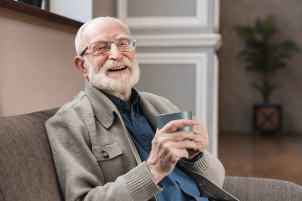 istock Delicious drink. Pleasant elderly man in spectacles sitting on the sofa and posing with a cup of coffee while smiling at the camera. Laughing elderly man sit relax on couch in living room drinking tea 1313904508