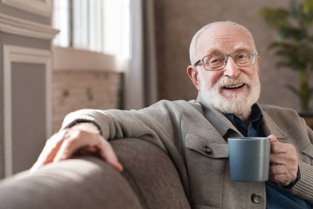 retrato recortado de un anciano anciano bebiendo café caliente sentado en el sofá interior. hombre mayor en gafas disfrutando de té caliente relajarse en la sala de estar - sofa men adult aging process fotografías e imágenes de stock