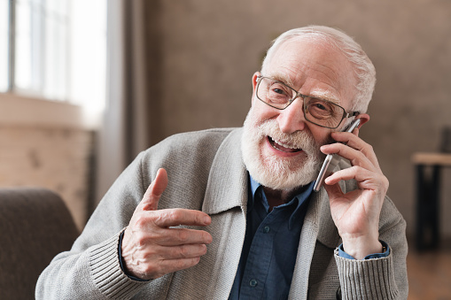 Overjoyed senior 80s grandfather sit on sofa at home laughing talking on modern cellphone, happy mature 70s man relaxing in living room speak have pleasant smartphone call, elderly and technology concept