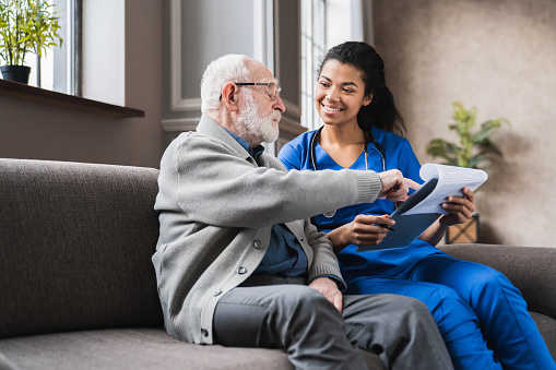 Female african-american professional doctor showing medical test result explaining prescription using clipboard visiting senior elderly old man patient at home sitting on sofa. Elderly people healthcare tech concept.