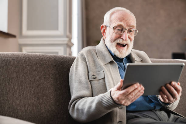 ソファの上にデジタルタブレットを使用して笑う年老いた祖父.肯定的な感情を持つ屋内で電子書籍を読む現代の高齢年金受給者 - one senior man only grandfather portrait old ストックフォトと画像