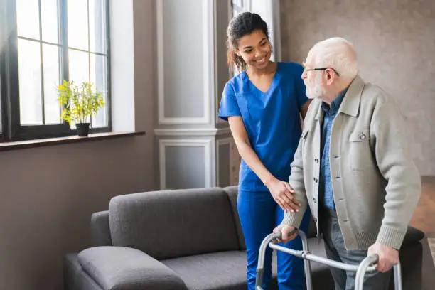 Photo of Portrait of an african young nurse helping old elderly disable man grandfather to walk using walker equipment in the bedroom. Senior patient of nursing home moving with walking frame and nurse support