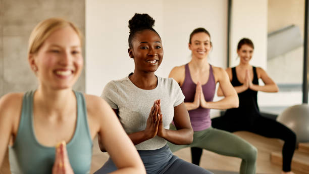 Happy African American female athlete practicing Yoga with group of women at health club. Group of athletic women with hands clasped doing Yoga exercises in health club. Focus is on black woman. yoga class photos stock pictures, royalty-free photos & images