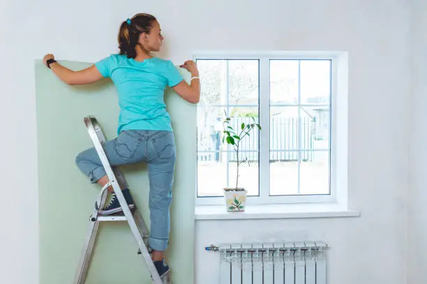 Woman in jeans and turquoise T-shirt standing on stepladder and holding piece of wallpaper. Home renovation and improvement.