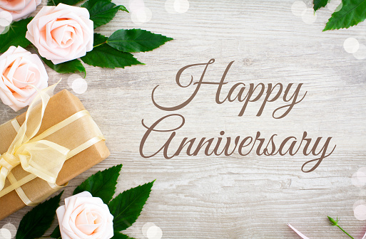 Happy Anniversary with pink roses and a gift with gold ribbon on a rustic wooden table.
Shot from above.