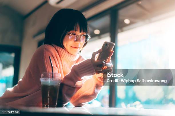 若い大人の幸せなアジアの女性は、ソーシャルメディアのアプリケーションのために携帯電話を使用して眼鏡を着用しています。