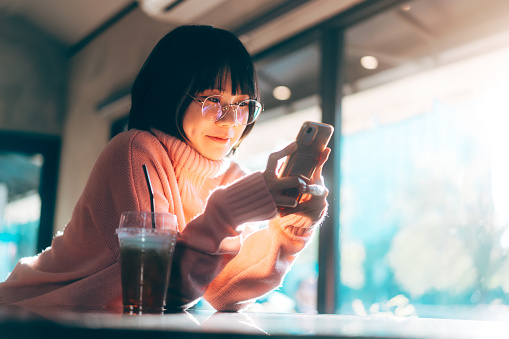 Young adult happy asian woman wear eyeglasses using mobile phone for social media application. Text online message at indoor cafe on day. Background with window and warm sunlight on winter season.
