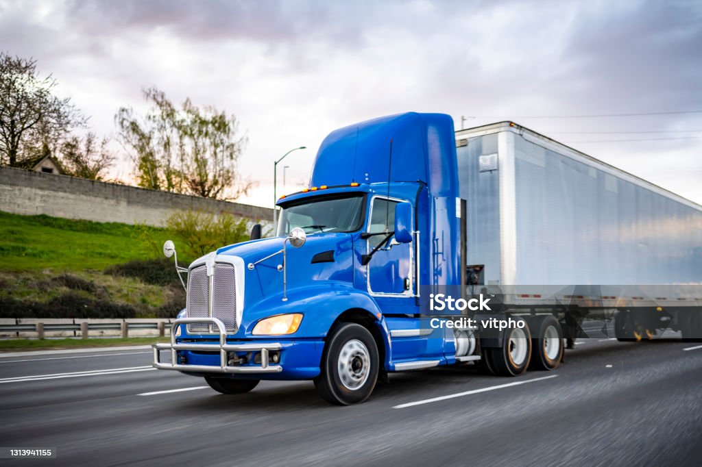 Powerful blue day cab big rig semi truck with dry van semi trailer driving on the wide road at twilight time Day cab blue big rig industrial semi trucks tractor with roof spoiler and turn on headlight transporting commercial cargo in dry van semi trailer running on the wide highway road at twilight time Semi-Truck Stock Photo