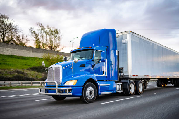 Powerful blue day cab big rig semi truck with dry van semi trailer driving on the wide road at twilight time Day cab blue big rig industrial semi trucks tractor with roof spoiler and turn on headlight transporting commercial cargo in dry van semi trailer running on the wide highway road at twilight time haulage stock pictures, royalty-free photos & images