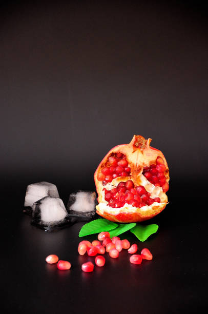 A pomegranate, a ripe fruit with leaves broken and lying on a black background next to seeds and cubes of melted ice. Close up shot.