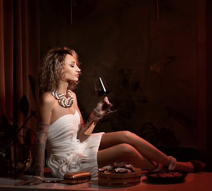 Portrait of rich arrogant blonde curly hair woman in white strapless dress and gloves sitting on table served with oysters and drinking wine over dark background