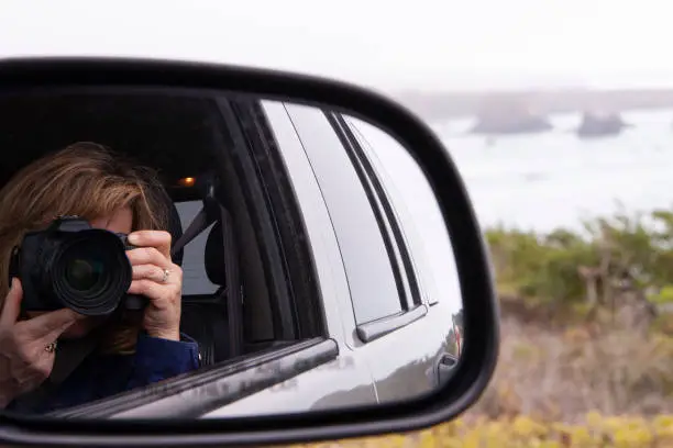 Woman holding a 35mm camera up to face taking a picture of self in rearview mirror while also seeing the ocean in the background