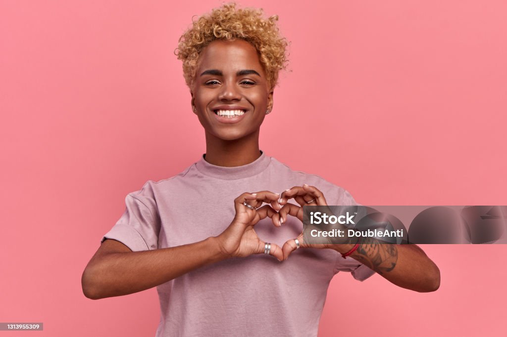 Portrait of happy african woman confesses in love, shows heart gesture, smiles broadly, has romantic feelings, seeks lonely hearts, feels passion, dressed in casual lilac t shirt, over pink background Heart Shape Stock Photo