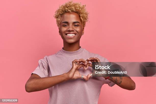 Portrait Of Happy African Woman Confesses In Love Shows Heart Gesture Smiles Broadly Has Romantic Feelings Seeks Lonely Hearts Feels Passion Dressed In Casual Lilac T Shirt Over Pink Background Stock Photo - Download Image Now