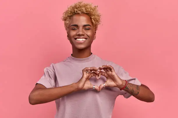 Portrait of happy african woman confesses in love, shows heart gesture, smiles broadly, has romantic feelings, seeks lonely hearts, feels passion, dressed in casual lilac t shirt, over pink background