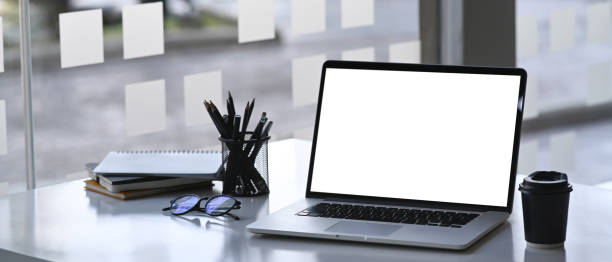 istock Mock up laptop computer with empty screen, coffee cup, notebook and pencil holder on white desk near window. 1313957298