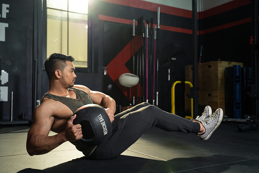 Working out at GYM concept. Young Asian handsome man working out his abdominal muscles by holding the medicine twist left and right sitting in the modern loft gym. Health and fitness concept.