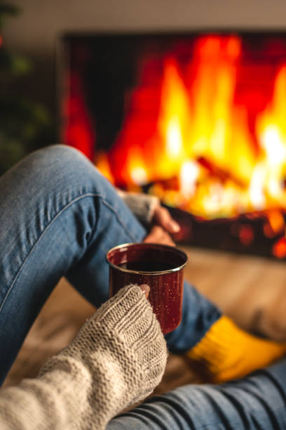 Girl in a sweater and socks is sitting next to the fireplace with cup of beverage. Concept of creating a cozy atmosphere stock photo