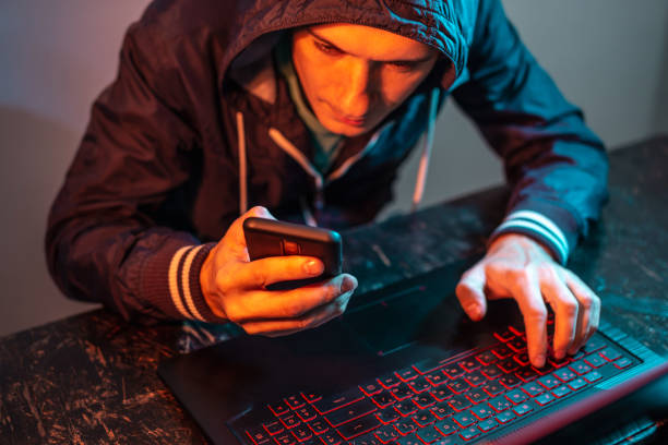 istock Hacker with phone is typing on a laptop keyboard in a dark room under a neon light. Cybercrime fraud and identity theft 1313967055